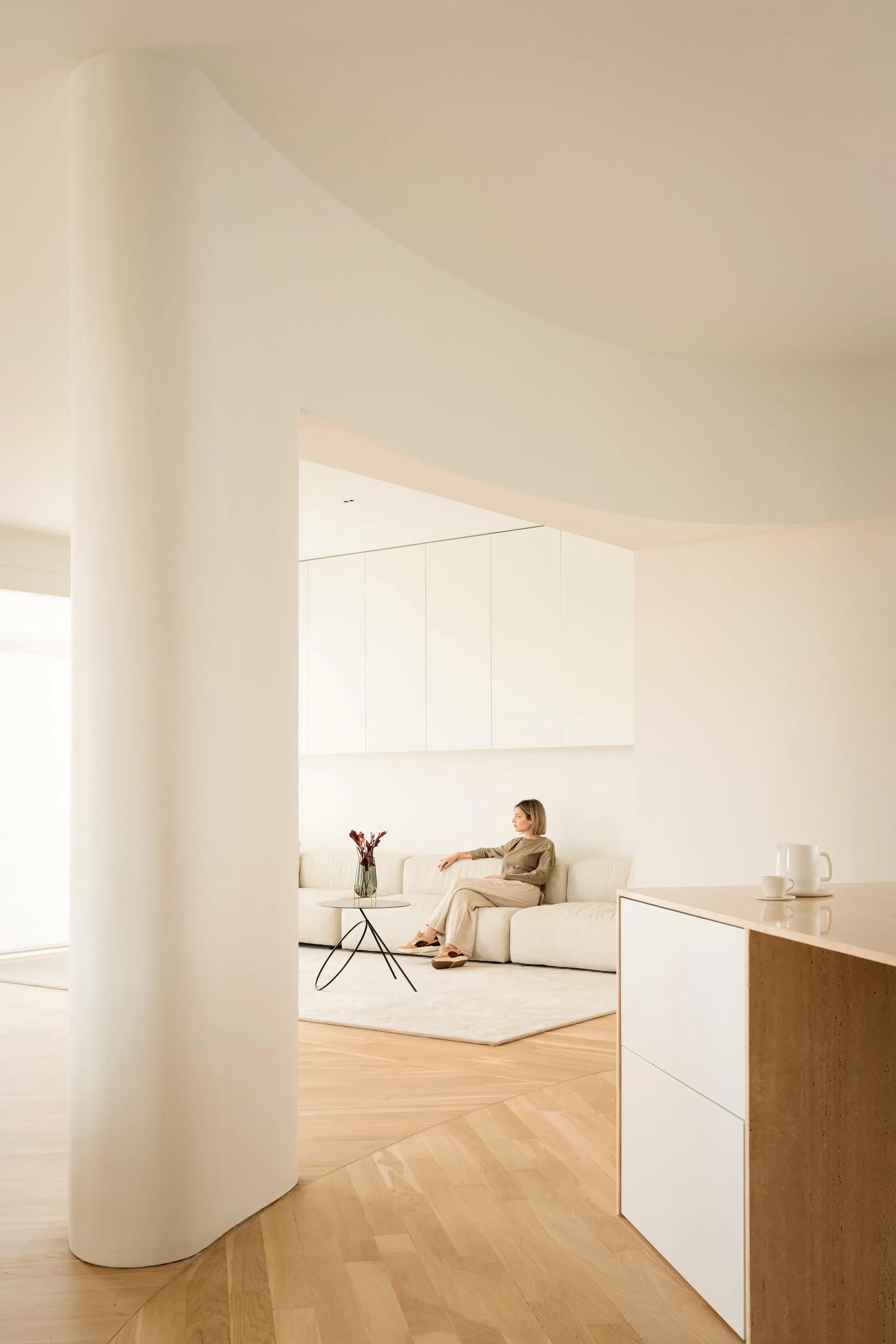 A woman sitting on a minimalist sofa in a modern living room with wooden flooring and white walls.