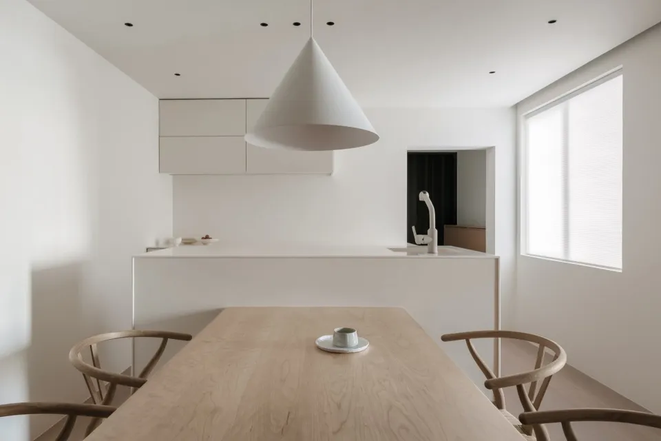 A minimalist residential kitchen with a white table, wooden chairs, and white pendant lighting.