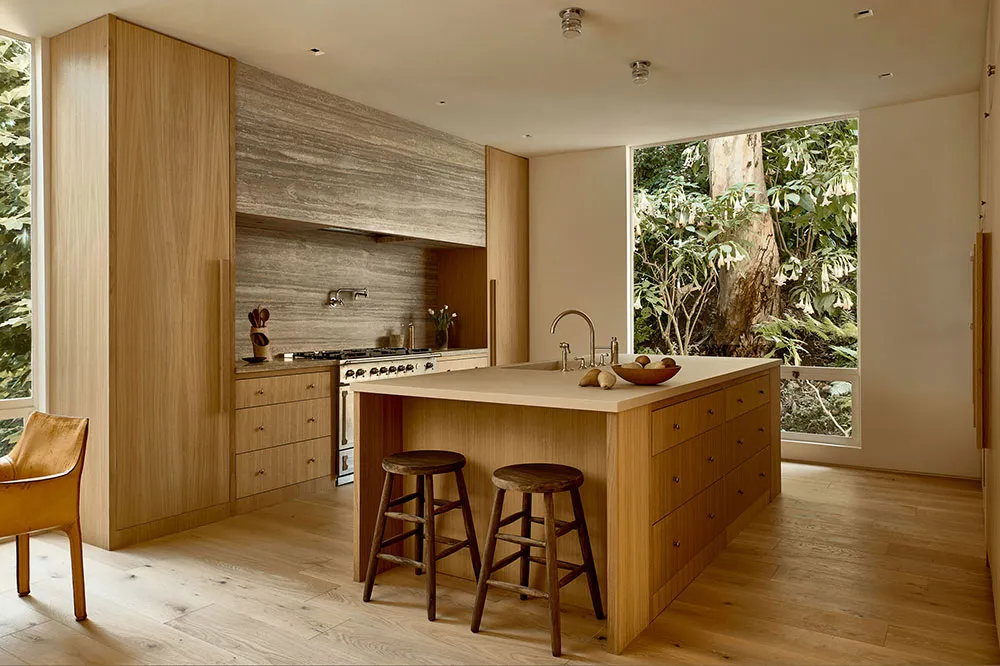 A modern kitchen with wooden cabinets, an island, and natural light from large windows overlooking greenery.