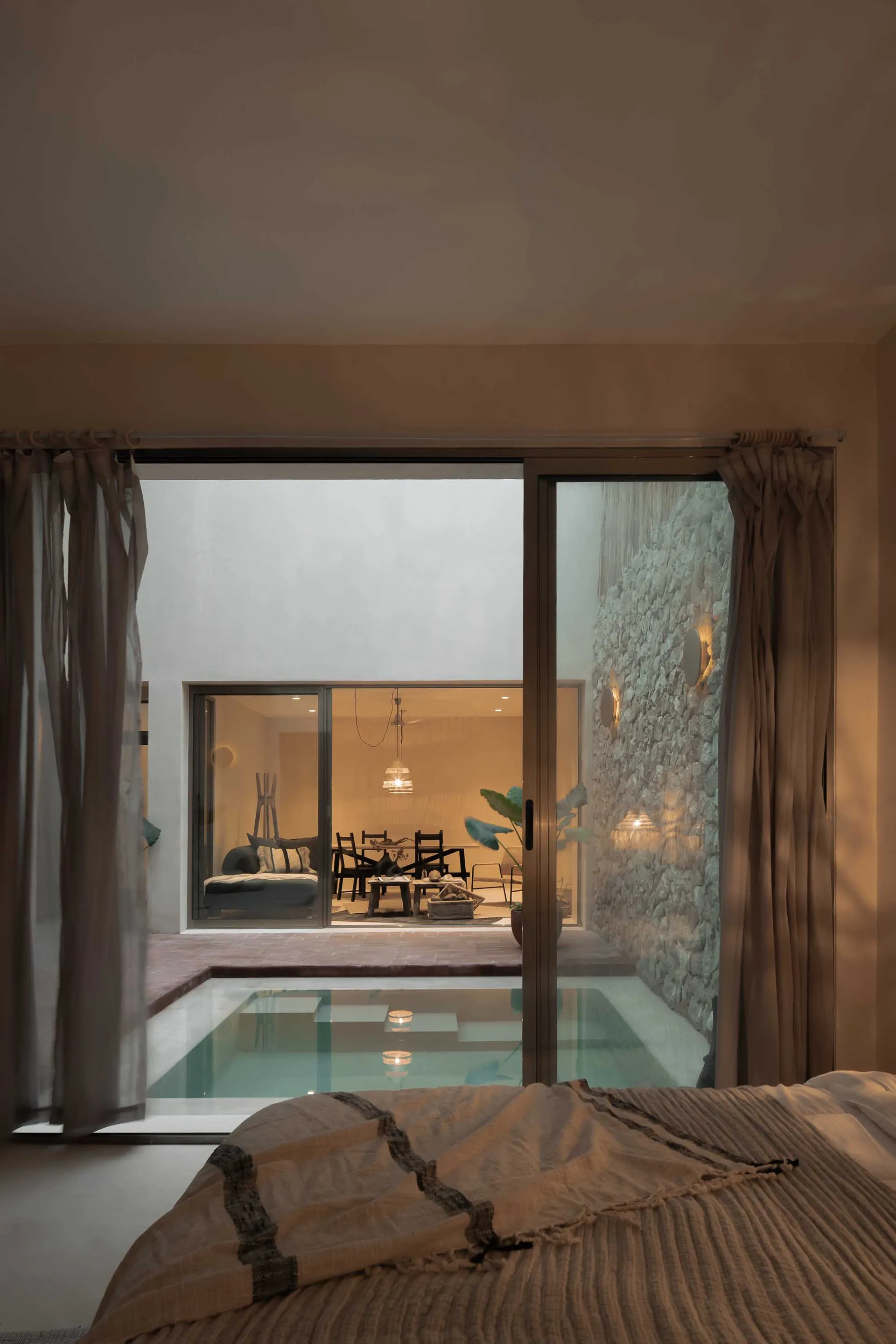 A modern bedroom with a concrete wall, a plunge pool, and a see-through glass wall that leads to a living area with a stone w