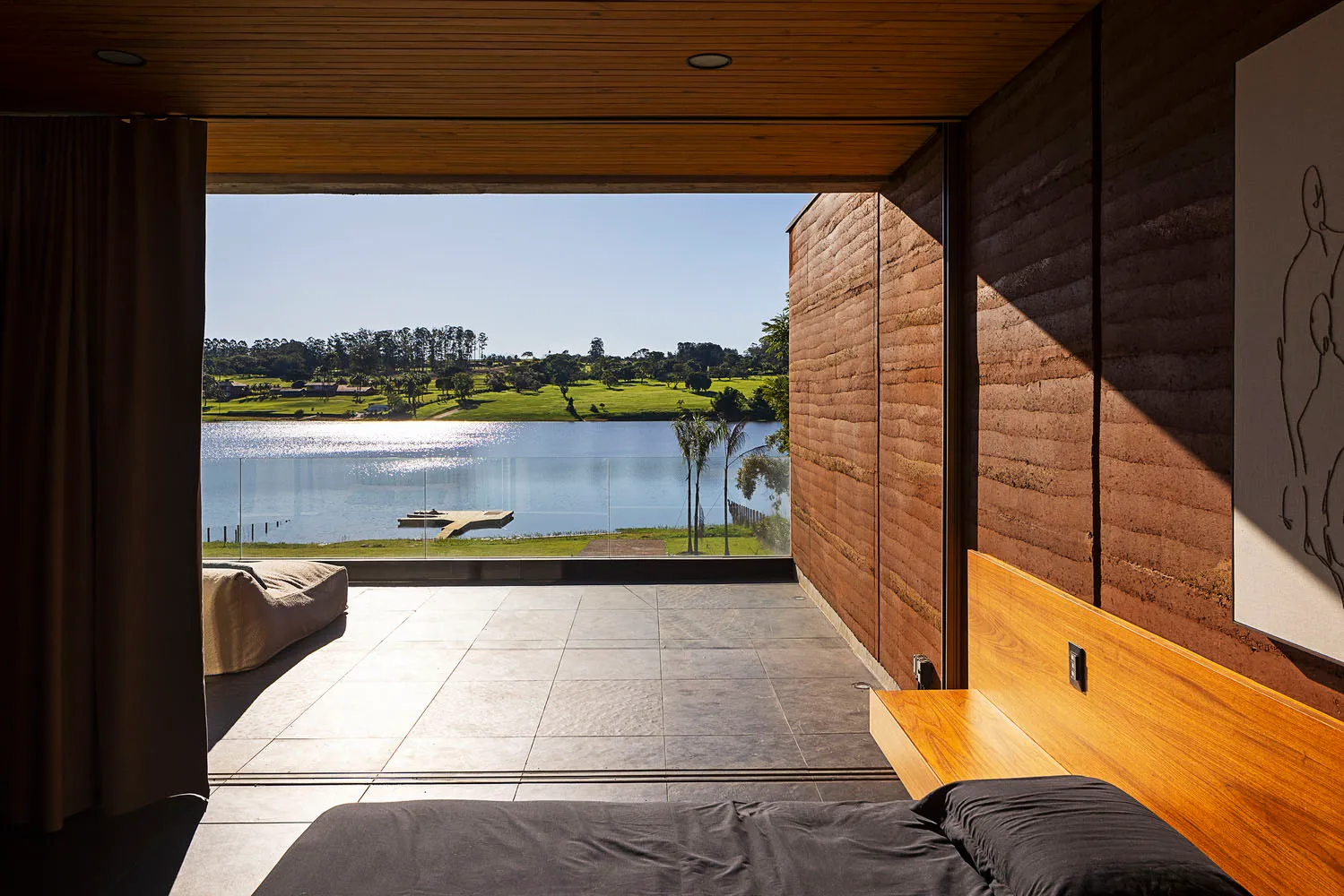 A minimalist bedroom featuring a black bedspread and a wooden headboard, with a view of a lake.