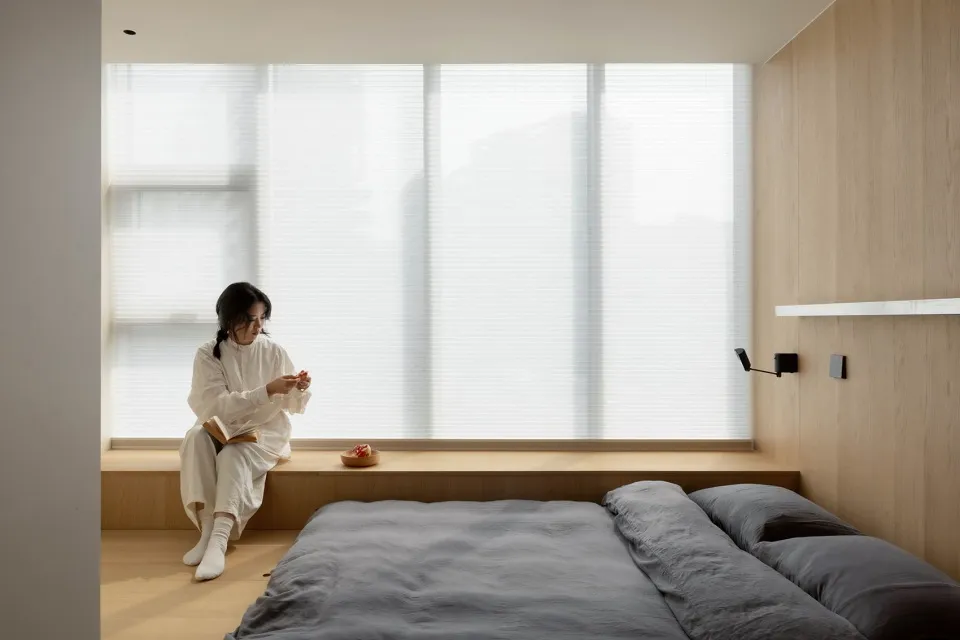 A minimalist bedroom featuring wooden elements with a person sitting by the window.