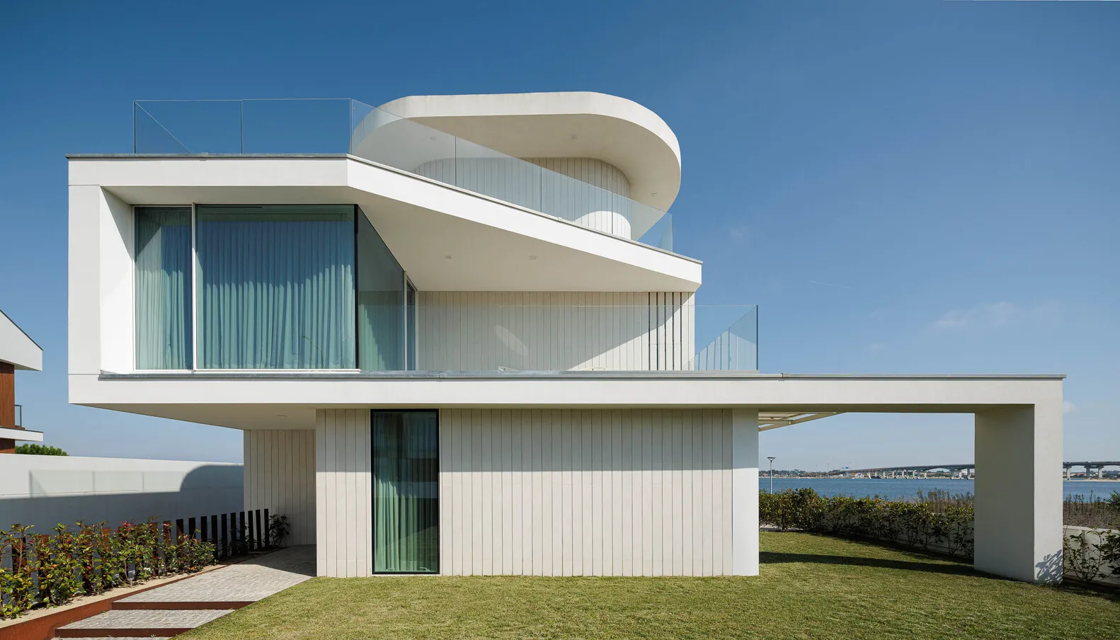 A modern house with white walls and large glass windows overlooking a body of water.
