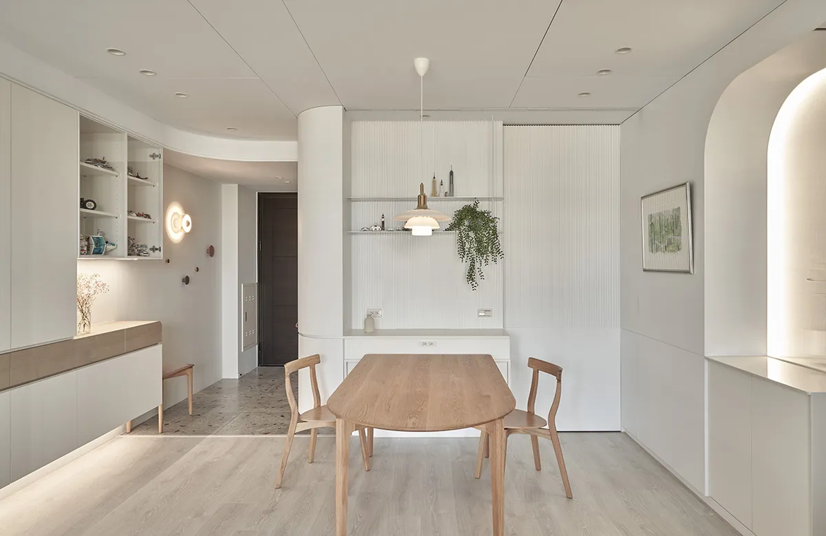 A minimalist dining room with a wooden table and chairs, white walls, and a hanging plant.