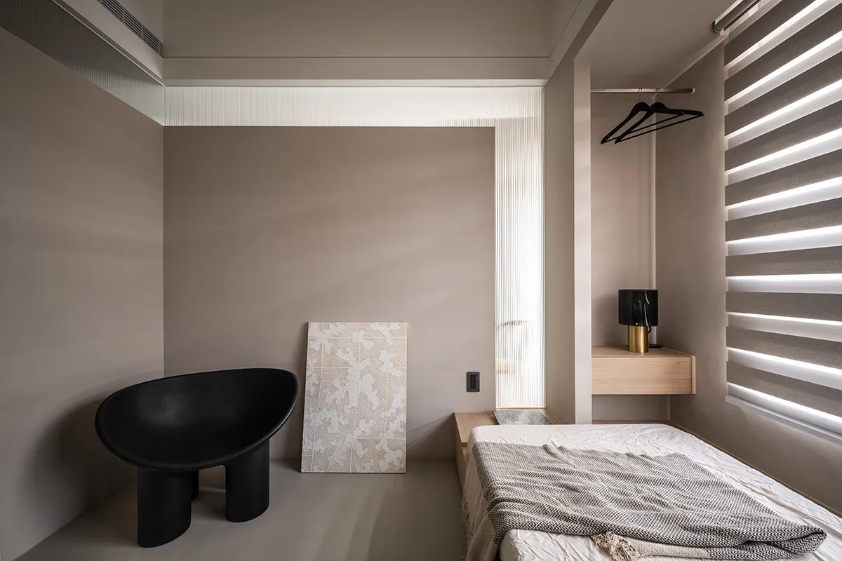 A minimalist bedroom featuring neutral-colored walls, a black chair, a bed with neutral bedding, and a wooden nightstand.