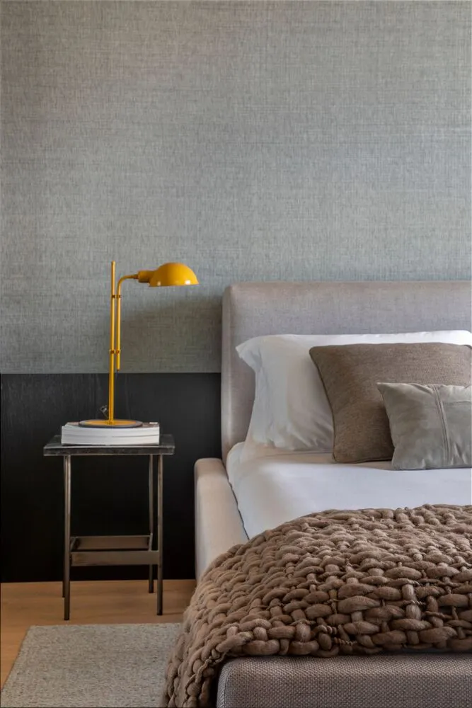 A minimalist bedroom featuring a grey upholstered bed with a taupe woven blanket, a yellow desk lamp, and a grey textured wal