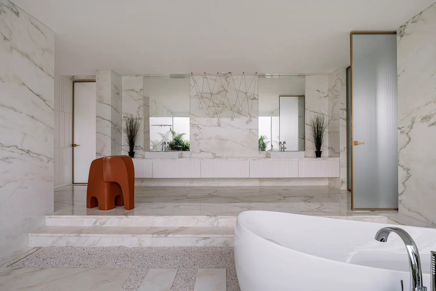 Modern bathroom interior featuring white marble walls, floor, and bathtub, with a red stool and potted plants.