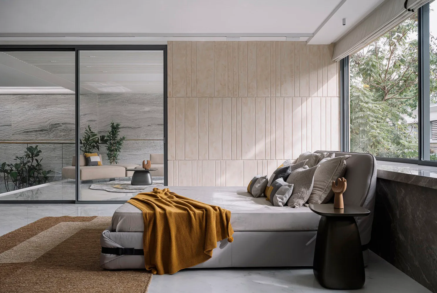 A modern bedroom featuring gray cushions and a yellow throw blanket on a gray sofa, with a view of green foliage outside.