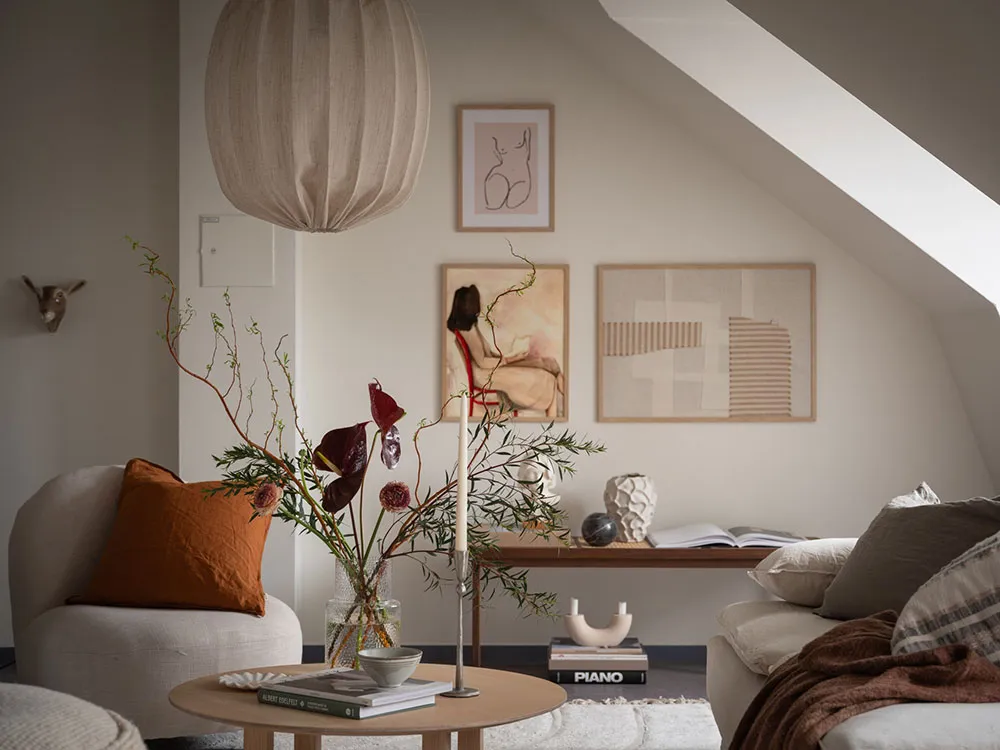 A cozy living room with neutral-toned decor, featuring a round coffee table, bookshelves, and floral arrangements.