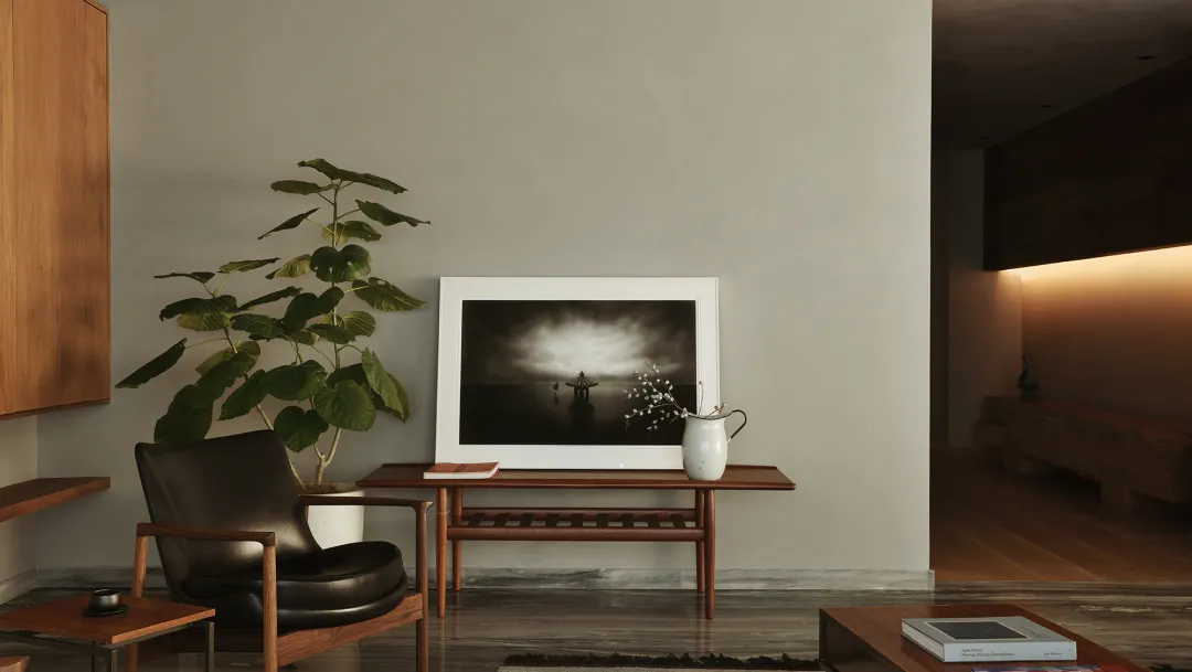 A minimalist living room with a black armchair, a table with a framed black-and-white photograph, and a potted plant on a pla
