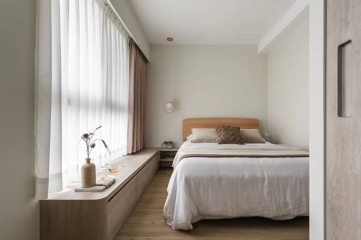 A minimalist bedroom with a wooden bed and wooden storage bench alongside the window, featuring sheer and light-colored curta