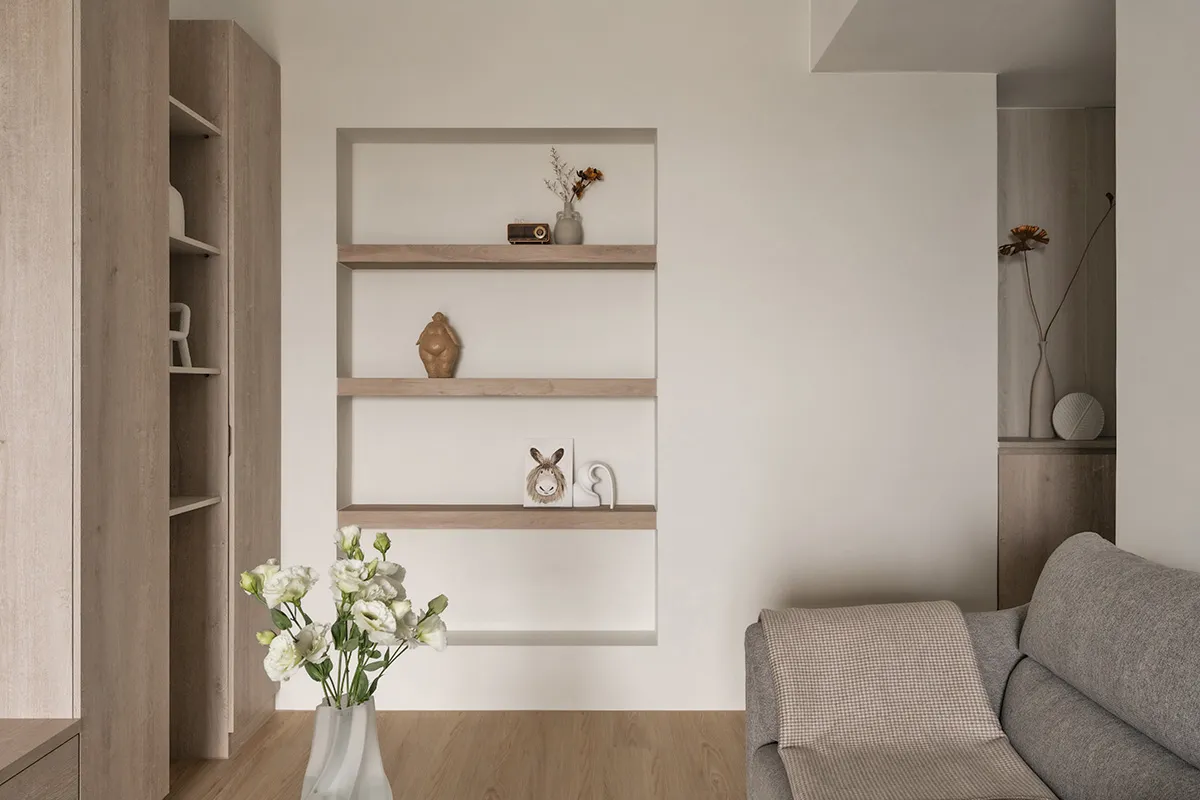 A minimalist living room featuring a gray couch, white flowers in a vase, and a floating shelf with decorative items on a neu