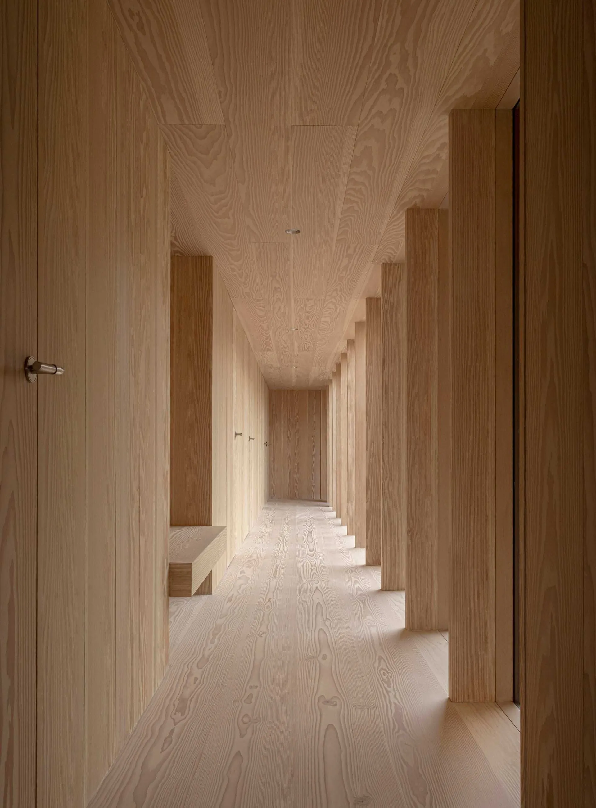 A minimalist corridor with wooden walls and ceiling, featuring long, narrow columns and a long, narrow bench.