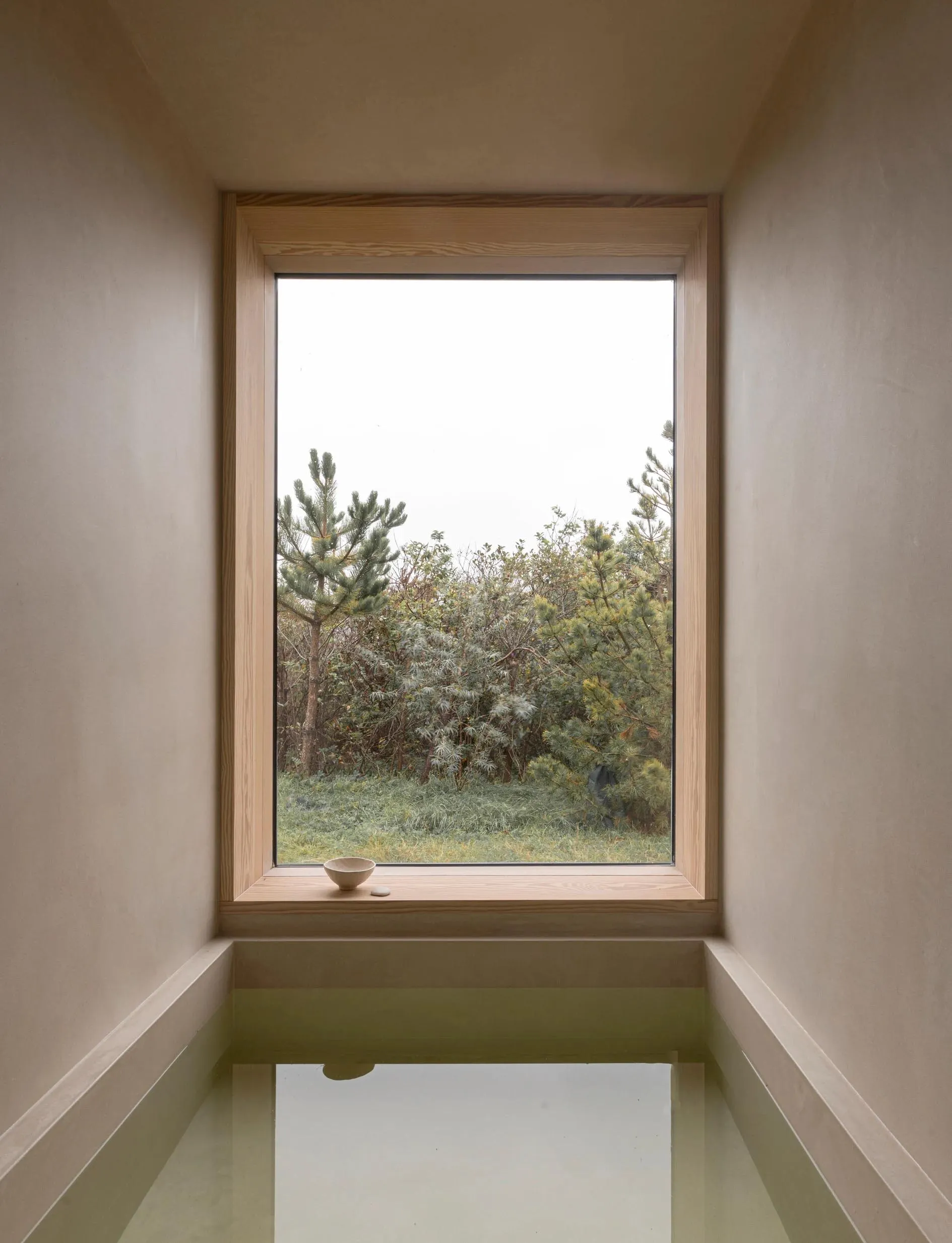 A minimalist bathroom with a glass window, wooden accents, and a concrete bathtub.