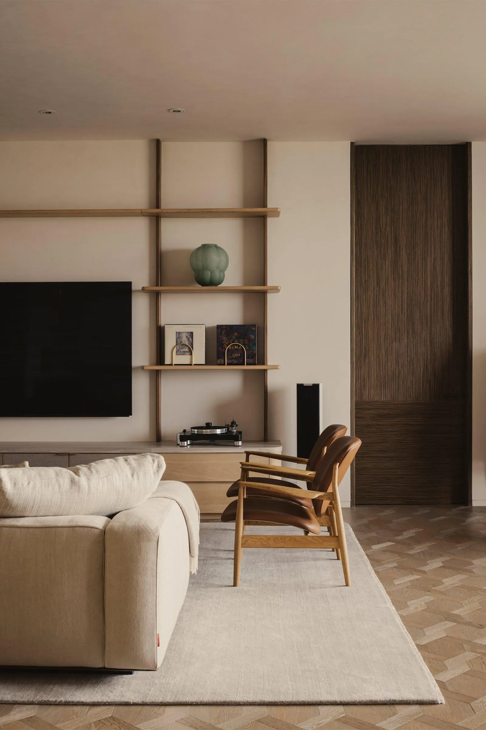 A modern minimalist living room with a beige sofa, brown armchairs, and a wooden shelf with books and a vase.