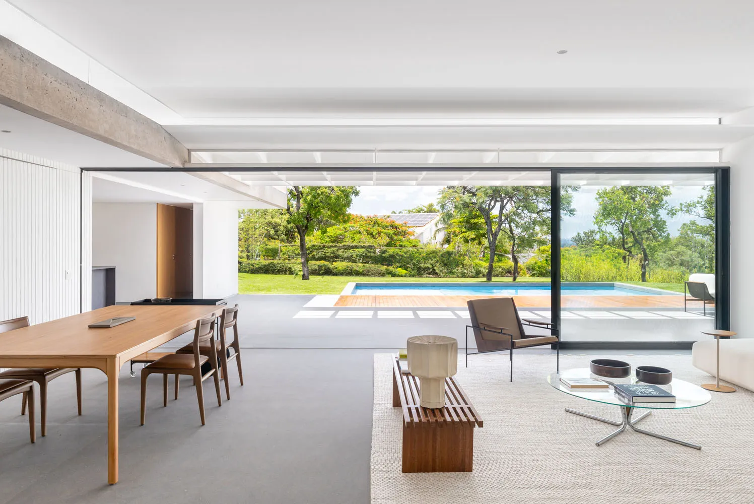 A modern living room featuring wooden tables, chairs, and a glass coffee table with a view of a pool and greenery.