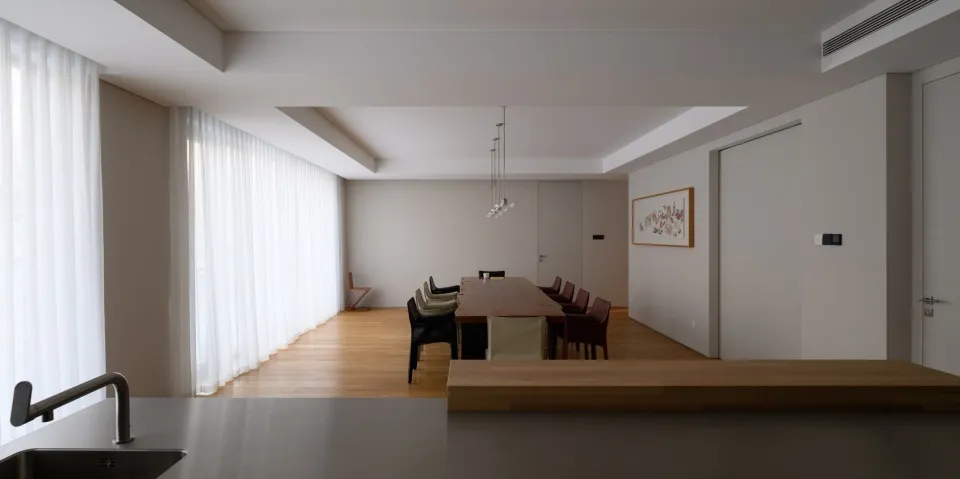 Modern minimalist dining room with a long wooden table, upholstered chairs, and pendant lighting.