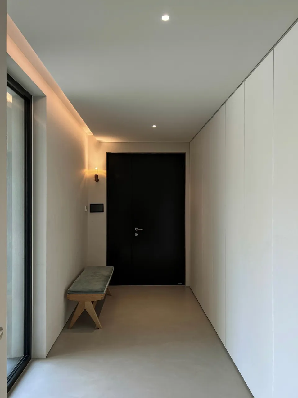 Minimalist interior design featuring a bench with a cushion and a black door in a concrete hallway
