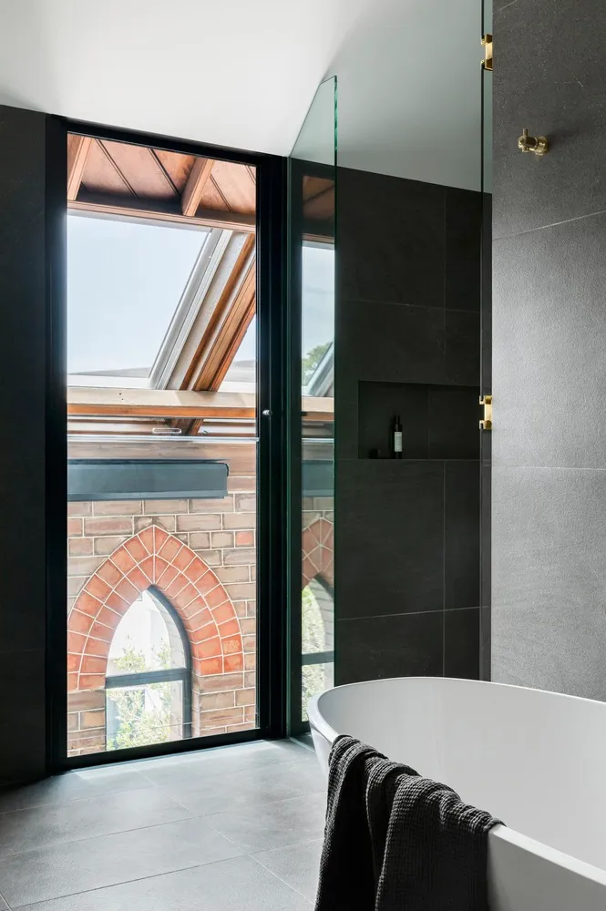 A modern bathroom with dark grey tiles, a freestanding bathtub, and a window overlooking an architectural brick structure.