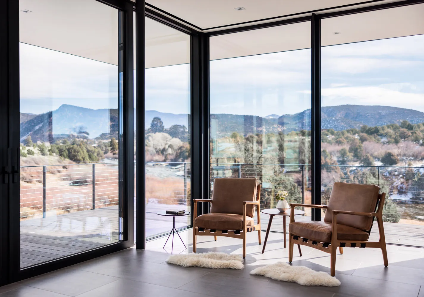 A modern living room with two brown leather chairs, a small side table, and a view of mountains through large windows.