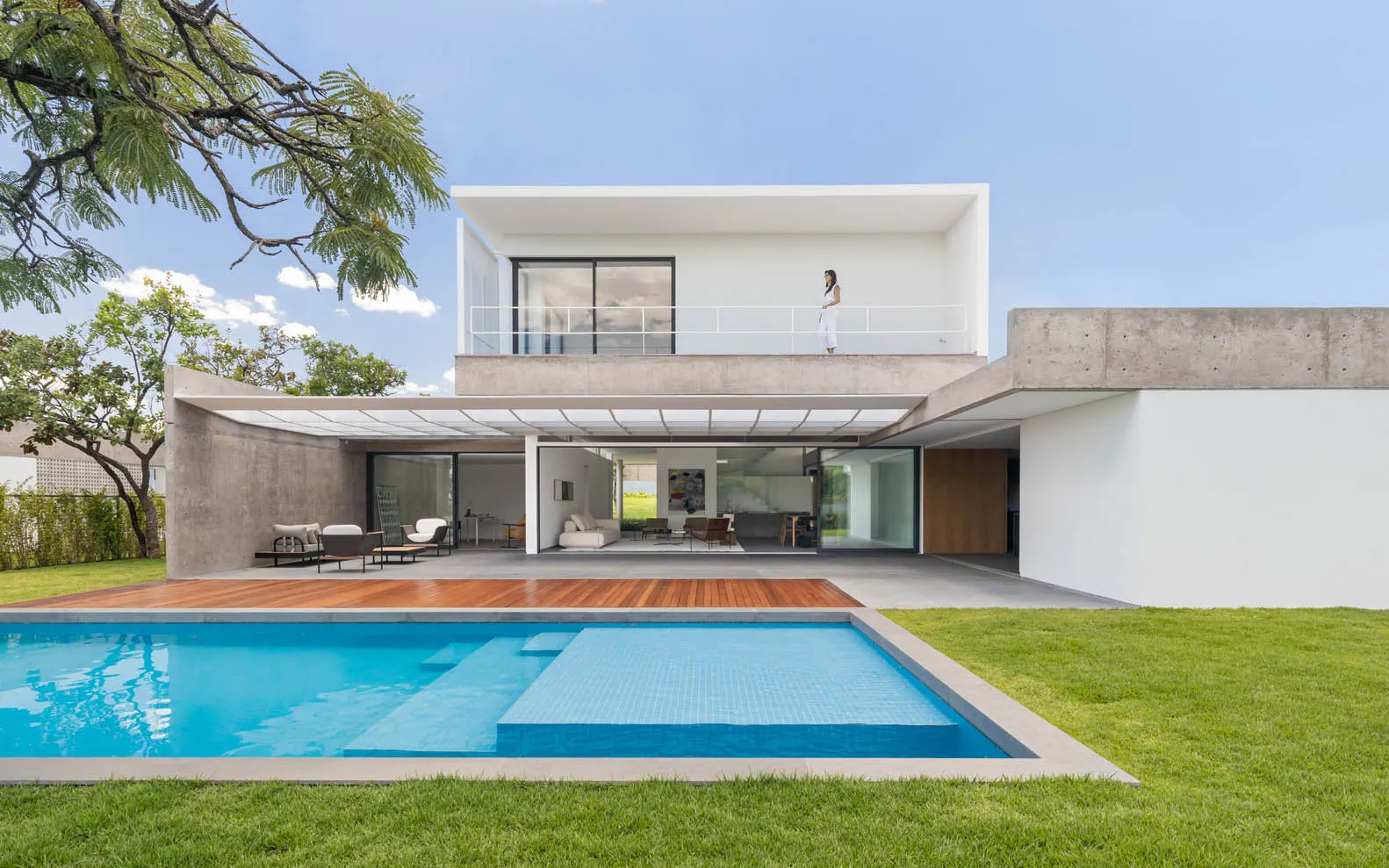 A modern house with a concrete exterior and a glass balcony overlooks a peaceful lawn and swimming pool.