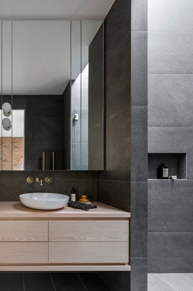 A modern bathroom with dark grey tiles, concrete sink, and brass faucet and wall.