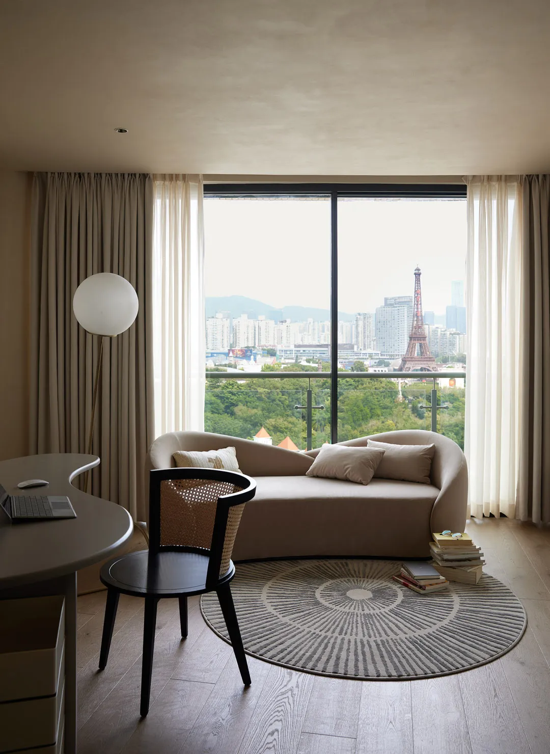 A modern residential interior featuring a beige couch with pillows, a black chair with a rattan weave, a round rug, a desk wi
