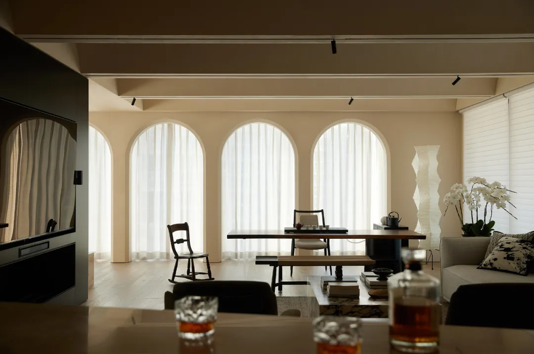 A modern living room with cream-colored walls and arched windows, featuring a wooden table, chairs, and a sofa with a glass a