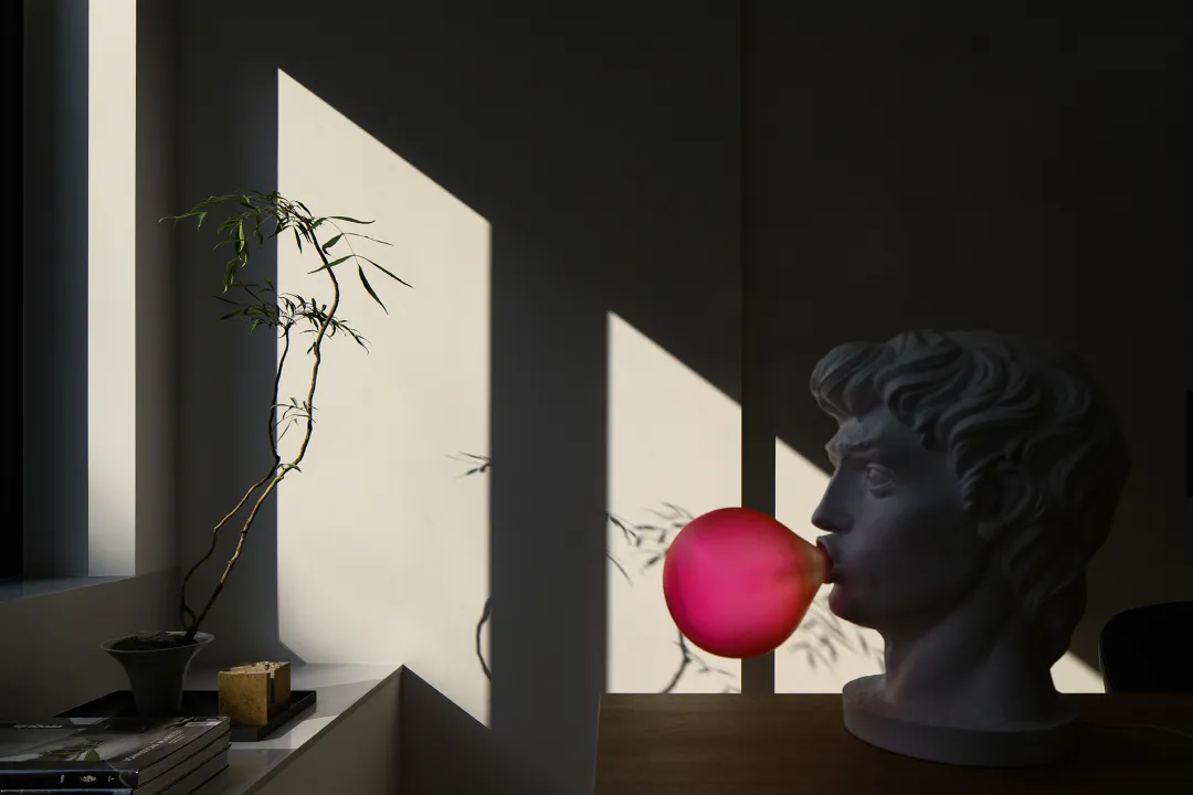 A ceramic sculpture blowing a bubble next to a window with sunlight streaming in, casting shadows on the wall.