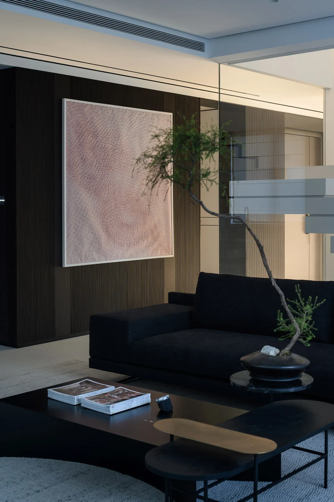A modern minimalistic living room featuring a dark sofa, a coffee table with books and a skateboard, and a bonsai tree.