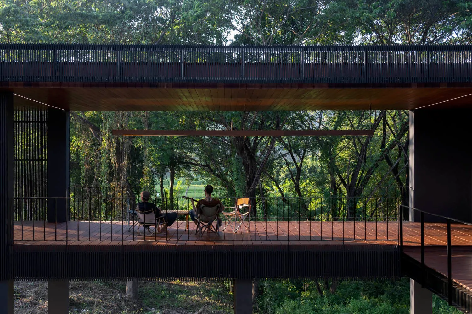 Three people sitting on a wooden deck surrounded by trees under modern industrial architecture.