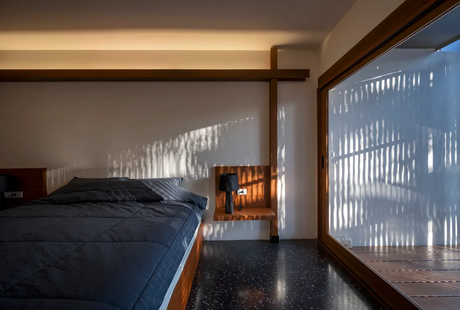 Modern minimalist bedroom with dark blue bedding, wooden headboard, and concrete floor.