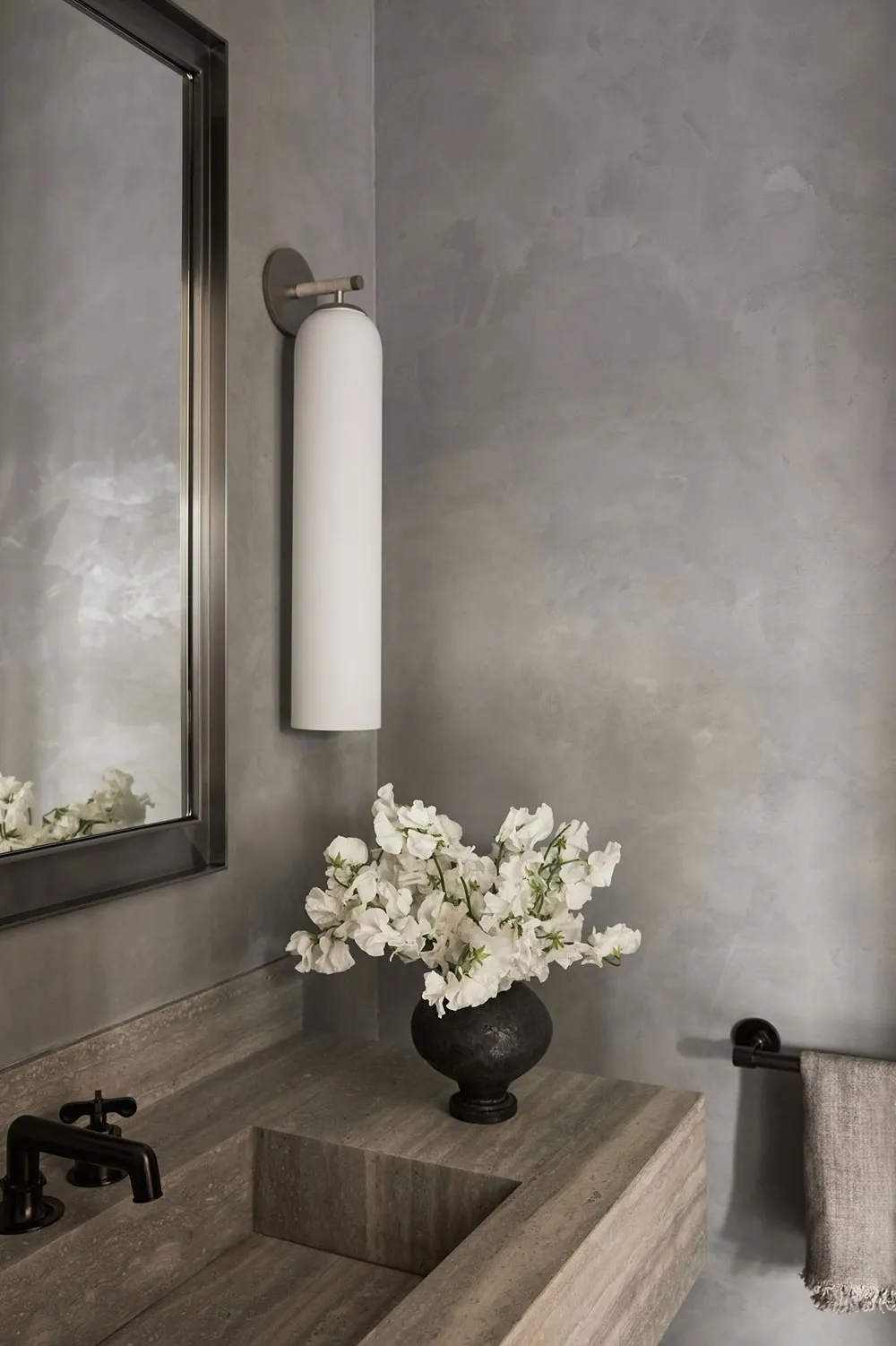 A modern, minimalist bathroom featuring concrete walls and a floating sink, with a vase of white flowers adding a touch of na