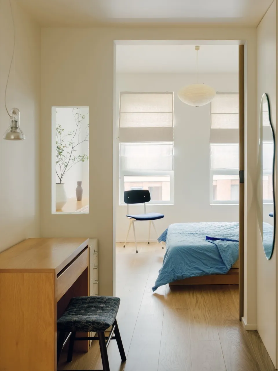 A modern, minimalist bedroom featuring hardwood flooring, a blue bedspread, and a dark blue upholstered stool.