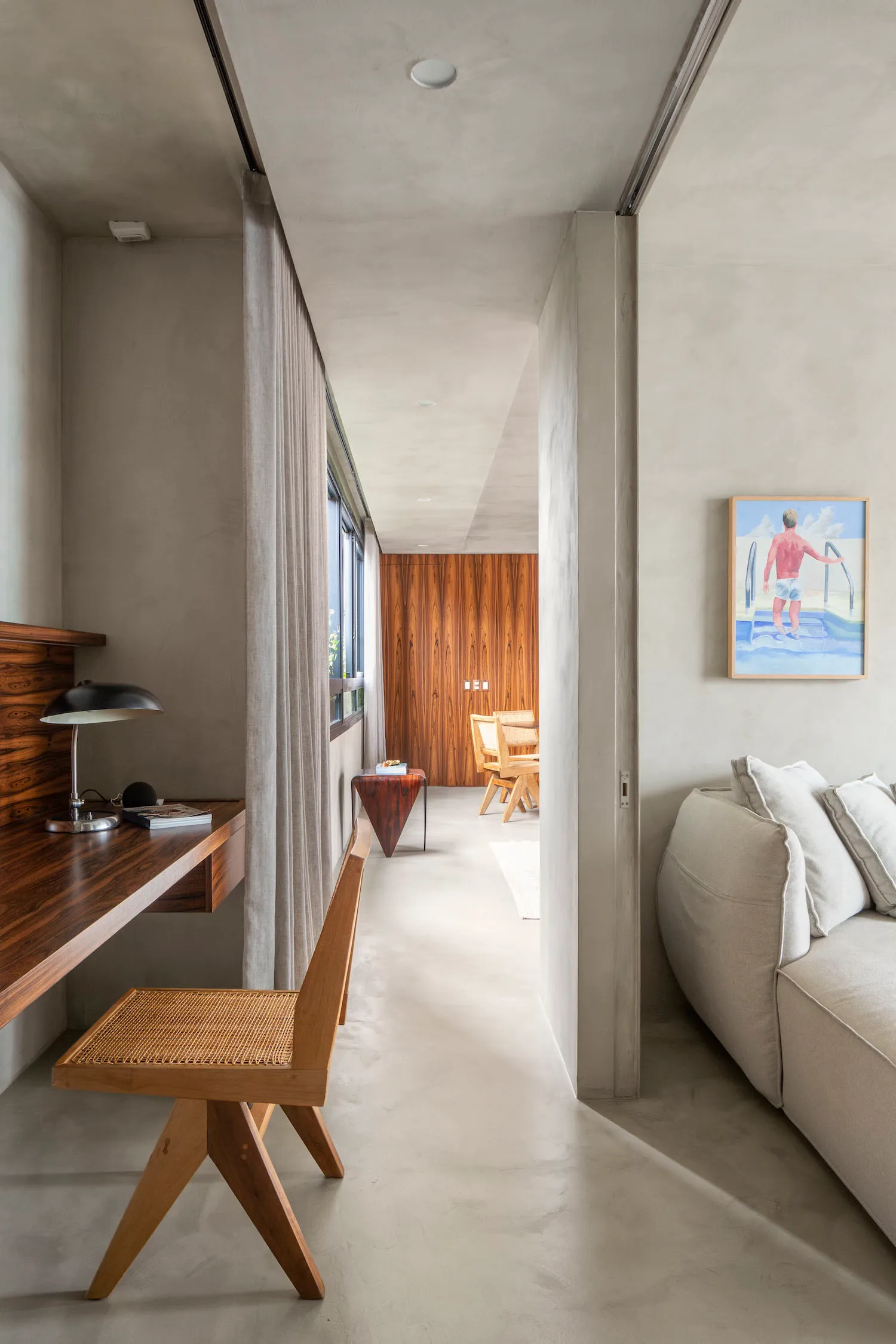 A modern minimalist interior space with concrete walls and floors, featuring a desk, chair, sofa, and artwork.
