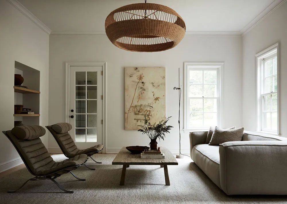 A minimalist living room with neutral-colored furniture, a large wicker chandelier, and a natural light-filled space.