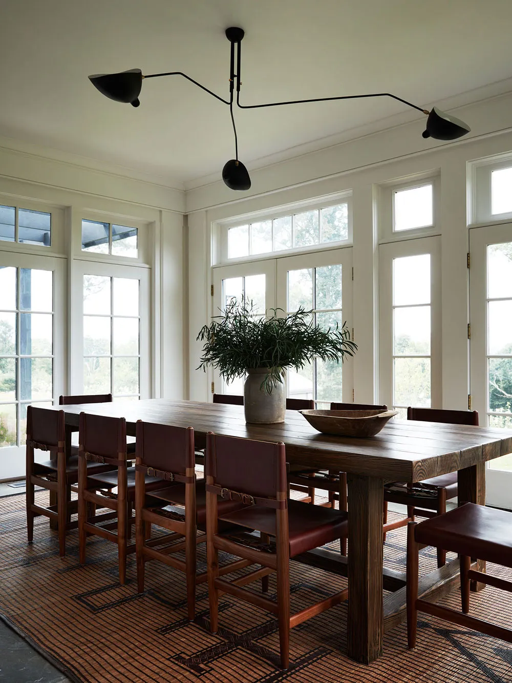 A minimalist dining room features a wooden table surrounded by brown leather chairs on a woven rug, with large windows and a