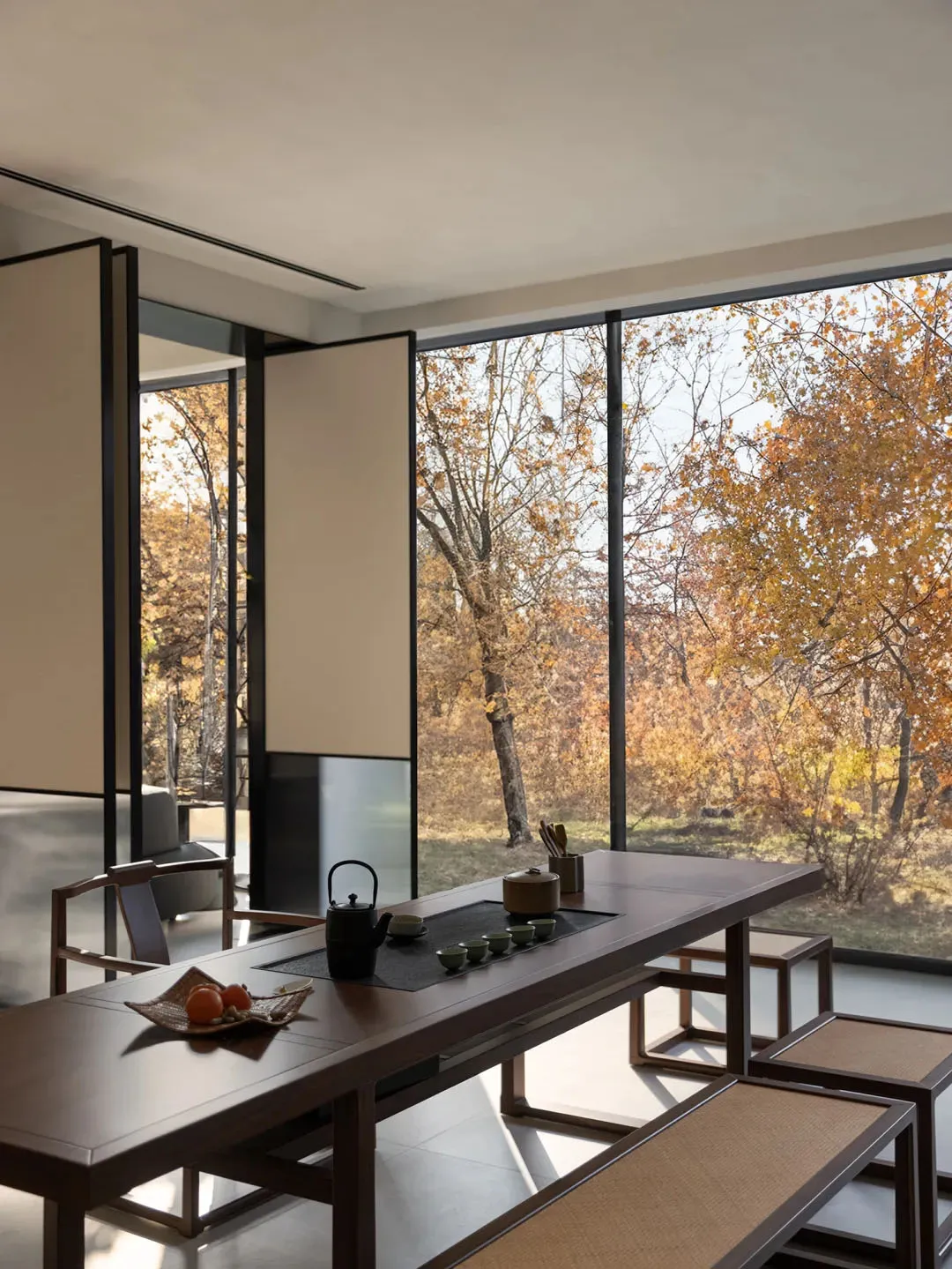 A modern coffee shop featuring a wooden counter with a tea set and autumn leaves outside the large windows.