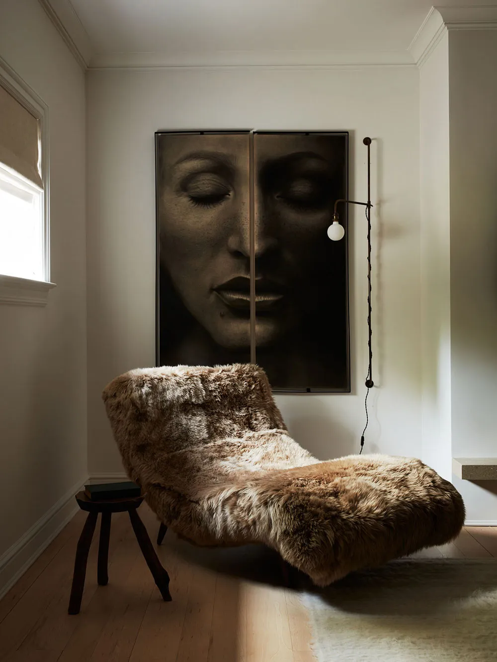 A modern living room with a fur blanket on a lounge chair, a large portrait of a face on the wall, and a small stool with a b