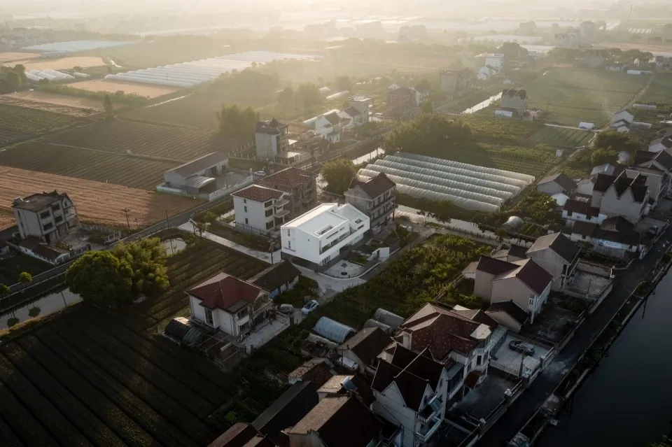 Aerial view of a village with houses, fields, and greenhouses under a hazy sky.