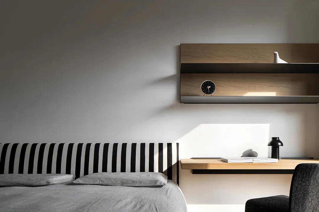 Modern minimalist bedroom featuring a bed with a black and white striped headboard and a light wood desk with a clock on it.