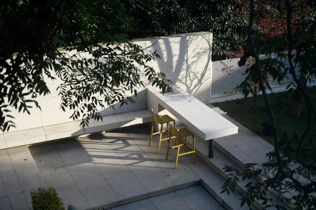 現代戶外空間,設有白色混凝土座椅區,包含桌椅,並被茂盛的綠植與樹木環繞