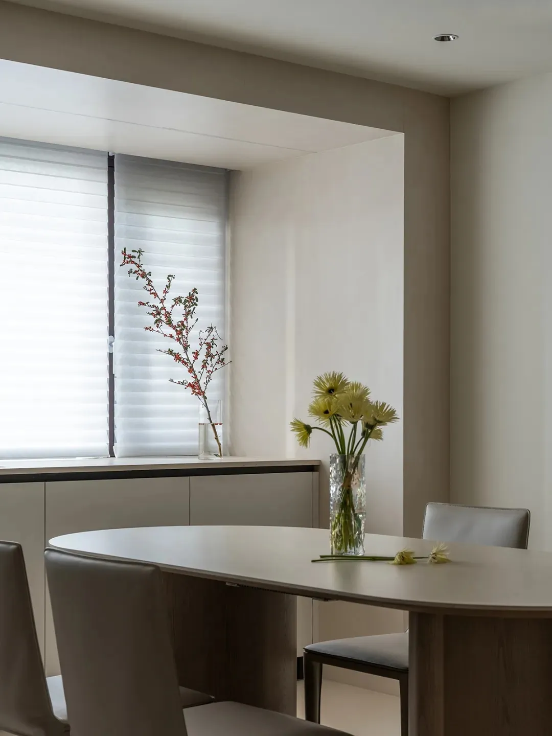Modern dining room with minimalist furniture and decor, including a round table, chairs, and fresh flowers in a vase by the w