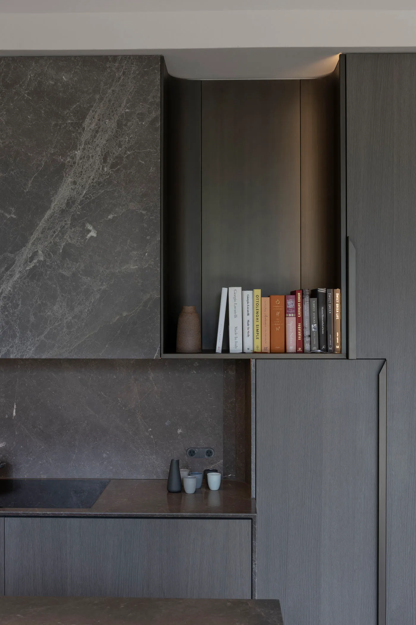 A modern residential kitchen area with grey marble surfaces and a bookshelf filled with cookbooks.