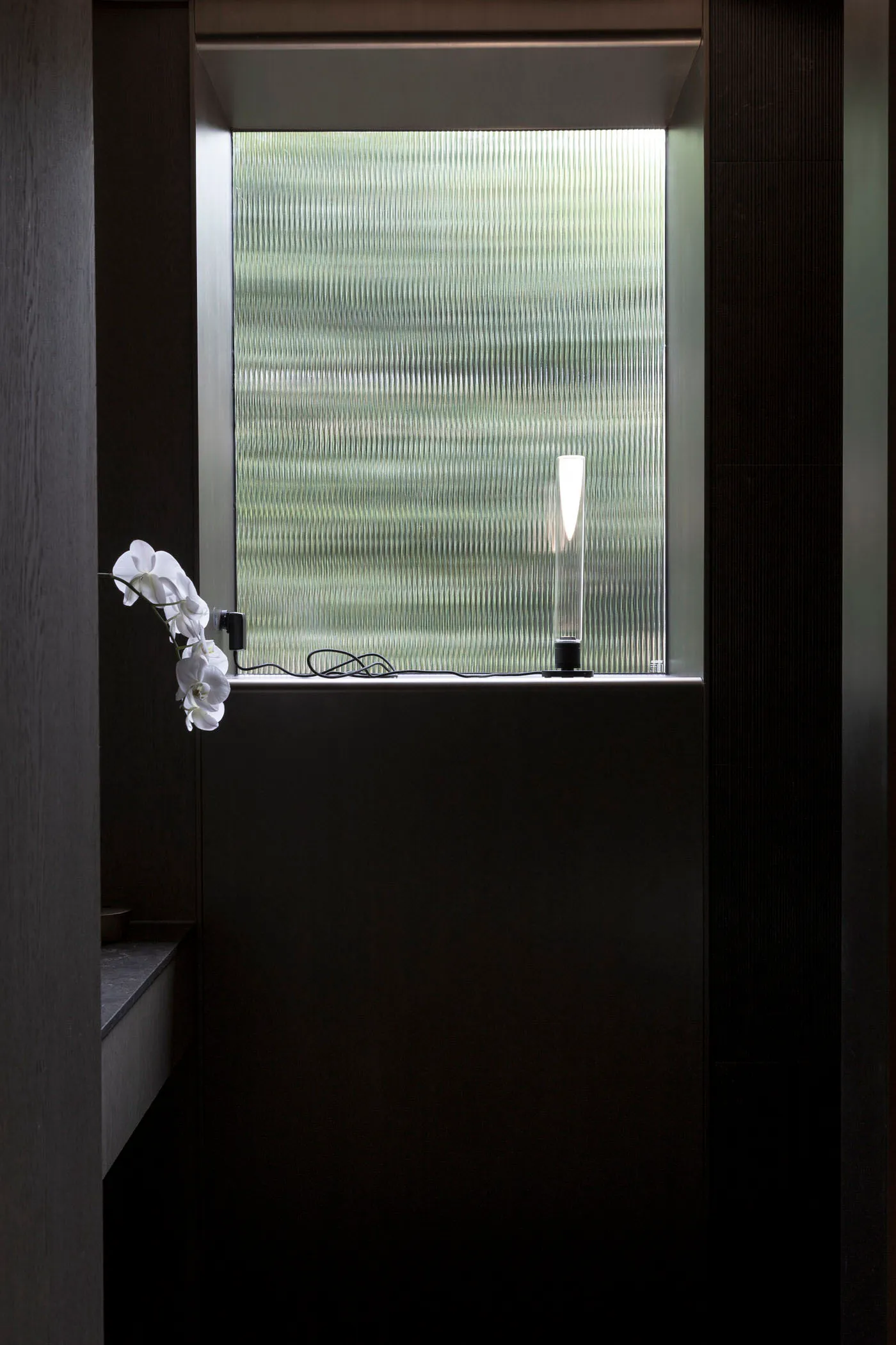 A minimalist bathroom featuring a frosted glass window with a sleek, modern lamp on the sill and a white orchid on the left s