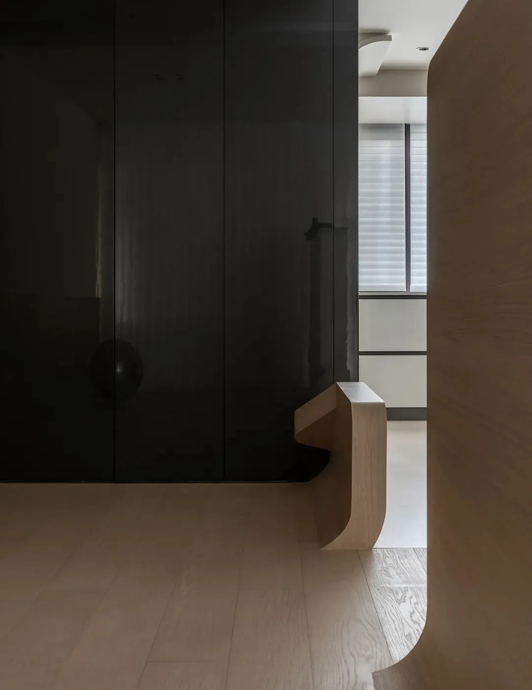 A minimalist entryway with a black glass wall and a wooden bench on white flooring