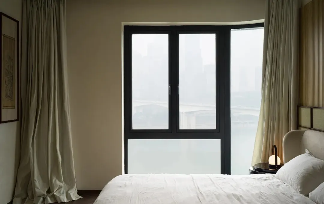 A modern bedroom with a bed covered in white bedding and white curtains hanging by the window.