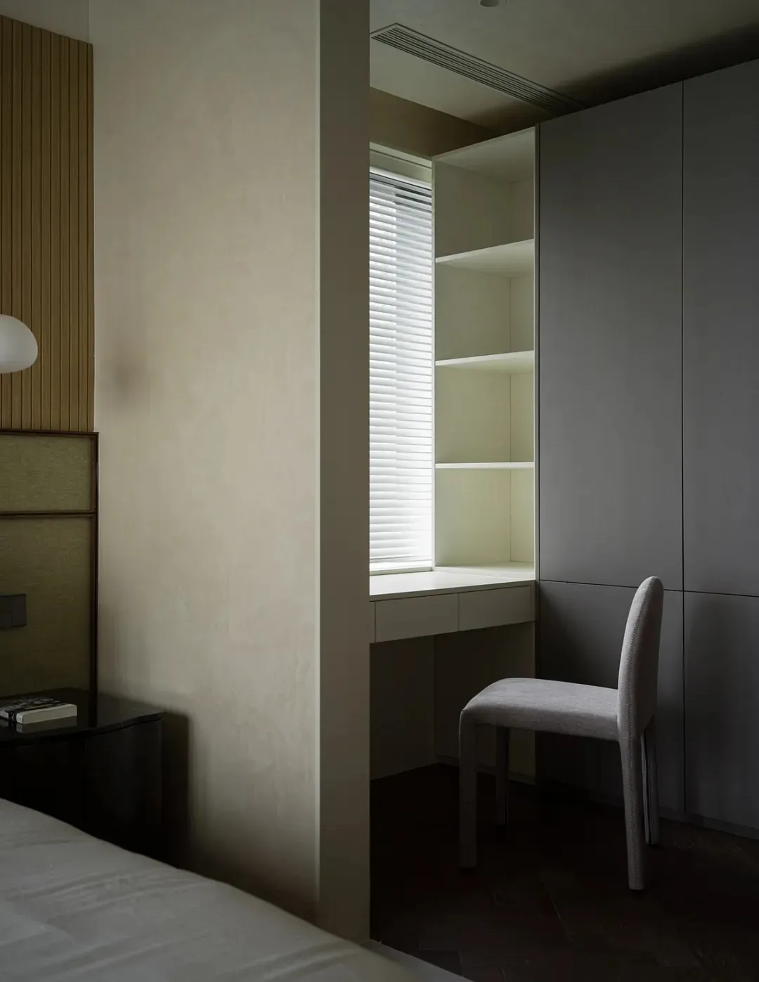 A modern minimalist bedroom featuring a bed with white sheets, a wooden headboard, a built-in desk with shelves, and a white