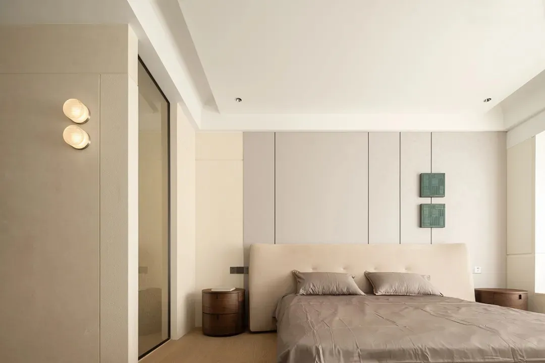 A minimalist bedroom featuring a bed with a beige headboard, grey linens, and a wooden floor, with a white and beige wall, an
