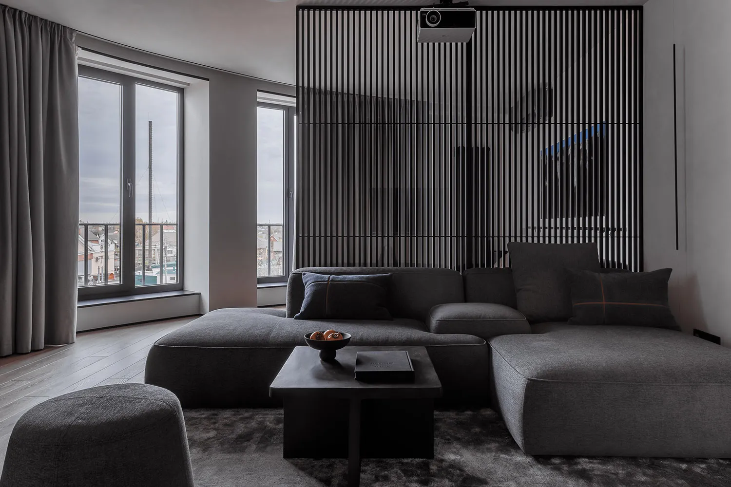 A modern, minimalist living room with a grey sofa, wooden floor, and large windows.